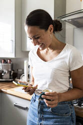 Lächelnde Frau, die ein Mobiltelefon benutzt und eine Kaffeetasse zu Hause in der Hand hält - WPEF07730
