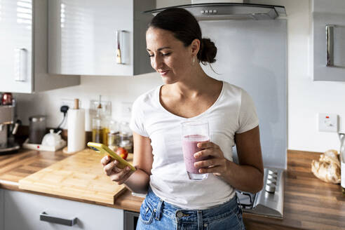 Lächelnde Frau, die ein Smartphone benutzt und ein Glas Milchshake zu Hause in der Hand hält - WPEF07740