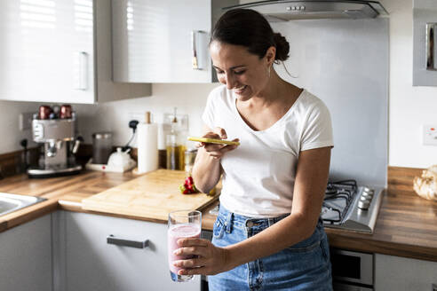 Glückliche Frau fotografiert Milchshake mit ihrem Smartphone in der Küche zu Hause - WPEF07741