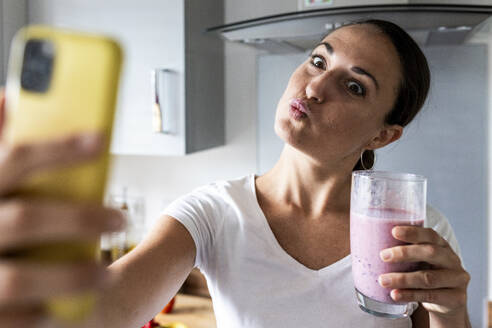 Woman puckering with glass of milkshake and taking selfie through smart phone - WPEF07743