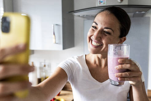 Happy woman clicking selfie with milkshake at home - WPEF07744