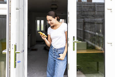 Smiling woman using smart phone in doorway at home - WPEF07761