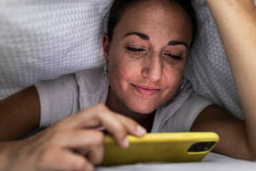 Lächelnde Frau, die ihr Smartphone unter einer Decke zu Hause benutzt - WPEF07783