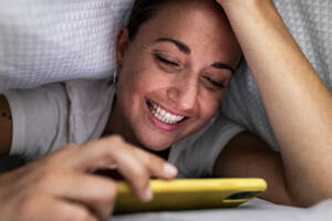 Glückliche Frau, die ihr Smartphone zu Hause unter der Bettdecke benutzt - WPEF07784