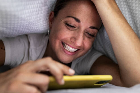 Happy woman using smart phone under blanket at home - WPEF07784
