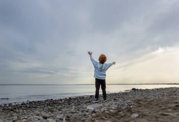 Carefree boy with arms raised standing at beach under sky - MBLF00010