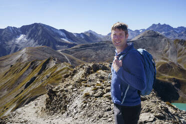 Glücklicher Mann mit Rucksack beim Wandern in den Bergen an einem sonnigen Tag - NJAF00592