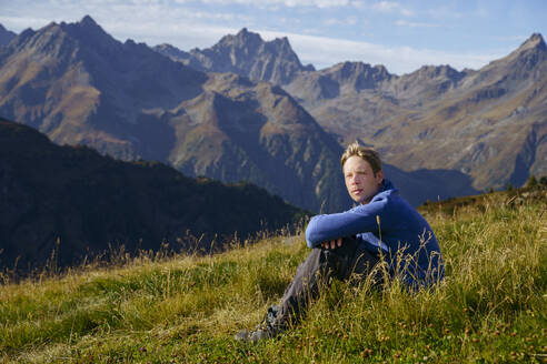 Mann sitzt auf einer Wiese in der Nähe eines Berges an einem sonnigen Tag - NJAF00593