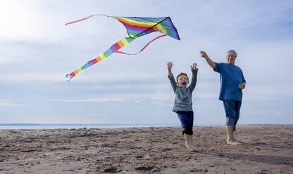 Happy boy flying multi colored kite with grandfather at beach - MBLF00021