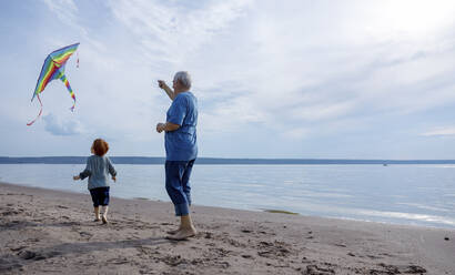 Älterer Mann lässt mit seinem Enkel am Strand einen bunten Drachen steigen - MBLF00022