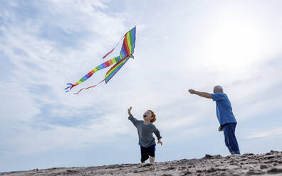Cheerful boy with grandfather flying kite under sky at beach - MBLF00023