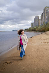 Young woman with hands in pockets walking on sand at waterfront - ANAF02304