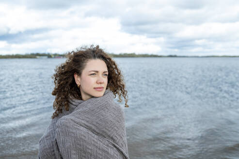 Nachdenkliche Frau mit lockigem Haar in Schal gewickelt am Wasser - ANAF02313