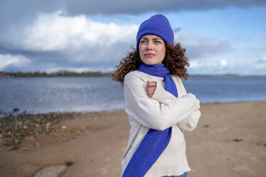 Contemplative woman wearing scarf and knit hat at beach - ANAF02325