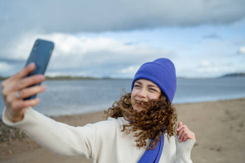 Smiling woman wearing knit hat taking selfie at beach - ANAF02326