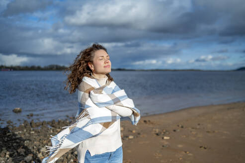 Happy woman with eyes closed wrapped in shawl at beach - ANAF02329