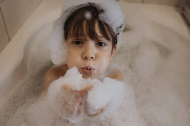 Cute boy blowing soap sud in bathroom at home - ANAF02334