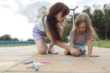 Woman teaching daughter to draw with chalk on tiled floor at park - LLUF01105