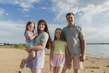 Smiling parents with daughters standing on sand at beach - LLUF01108