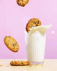 Flying oatmeal cookies near milk splashing in glass against pink colored background - FLMF01036