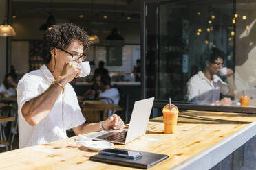 Young freelancer drinking coffee and sitting with laptop in cafe - PBTF00341