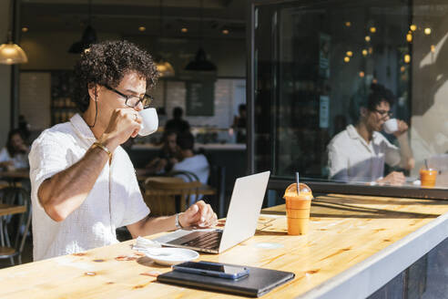 Junger Freiberufler trinkt Kaffee und sitzt mit Laptop in einem Cafe - PBTF00341