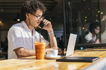 Junger Freiberufler, der in einem Café mit seinem Smartphone telefoniert und einen Laptop benutzt - PBTF00342