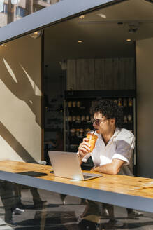 Junger Freiberufler trinkt Eiskaffee und sitzt mit Laptop in einem Café - PBTF00343