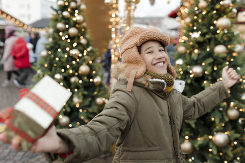 Fröhlicher Junge hält eine Geschenkbox und freut sich auf dem Weihnachtsmarkt - VIVF01105