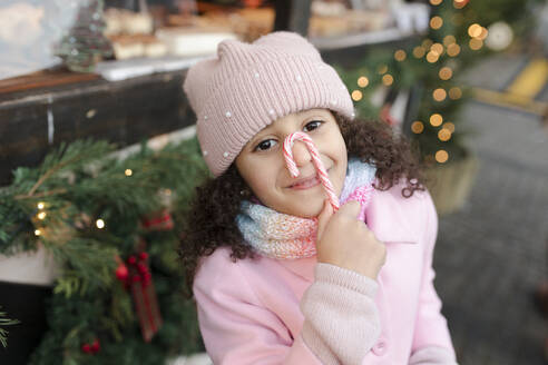 Smiling girl holding candy cane over face at Christmas market - VIVF01107