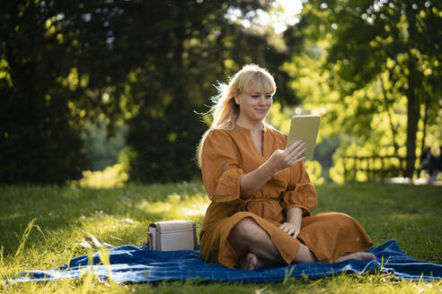 Smiling woman sitting on blanket and using tablet PC in park - JOSEF21465