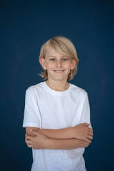 Happy boy standing with arms crossed against blue background - JOSEF21509