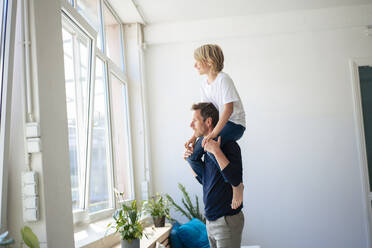 Father carrying son on shoulders and looking through window at home - JOSEF21535