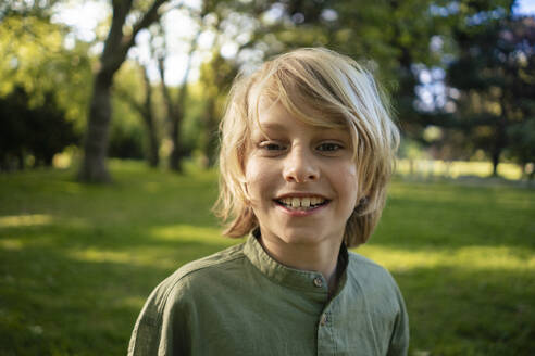 Smiling blond boy in front of trees at park - JOSEF21539