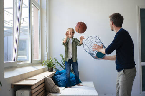 Happy boy playing basketball with father holding wastepaper basket - JOSEF21553