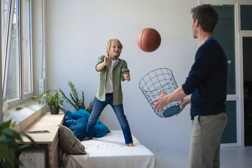 Happy boy throwing basketball with father holding wastepaper basket at home - JOSEF21556