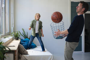 Happy boy with father catching basketball through wastepaper basket at home - JOSEF21557