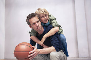 Boy leaning on father holding basketball in front of wall - JOSEF21565