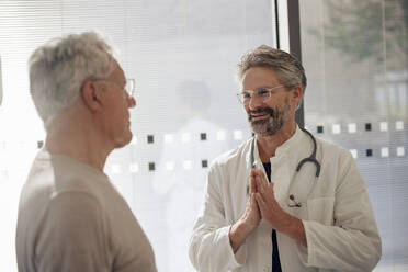 Smiling senior doctor wearing eyeglasses discussing with man in hospital - JOSEF21660