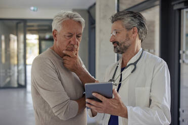 Senior doctor discussing with man over tablet PC in hospital corridor - JOSEF21671