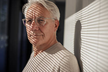 Smiling senior man wearing eyeglasses with sunlight over face near wall - JOSEF21686