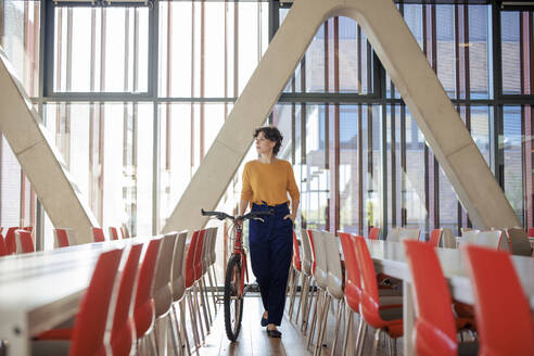 Young businesswoman walking with bicycle near chairs in office cafeteria - JOSEF21692