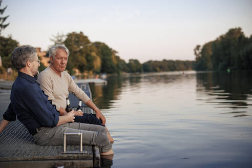 Senior friends sitting on pier with beer bottles near lake - JOSEF21713