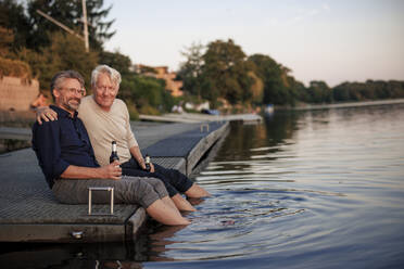 Smiling senior friends sitting on pier with beer bottles - JOSEF21714