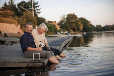 Happy senior friends sitting on jetty with beer bottles near lake - JOSEF21715