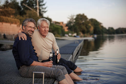 Smiling senior friends sitting with beer bottles on jetty - JOSEF21716