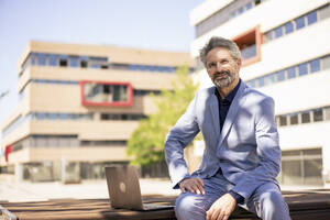 Lächelnder Geschäftsmann sitzt mit Laptop vor einem Gebäude - JOSEF21718