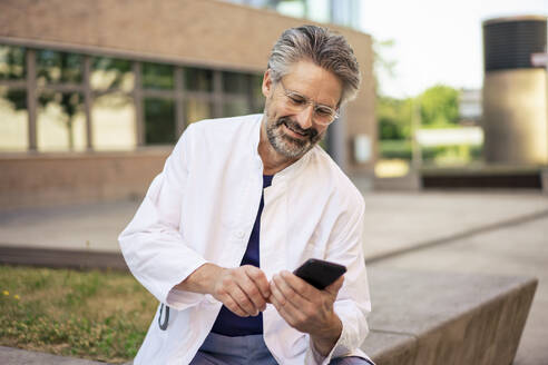 Smiling doctor using mobile phone in front of building - JOSEF21730