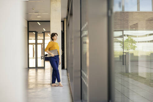 Contemplative businesswoman standing with laptop in office - JOSEF21742