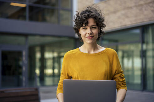 Lächelnde Geschäftsfrau mit Laptop vor einem Bürogebäude - JOSEF21747